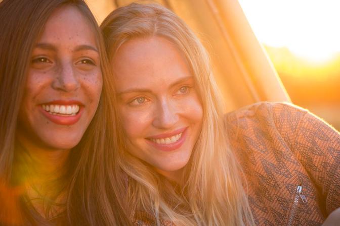 Two women with big smiles, embracing each other, both wearing casual, summery clothing, likely in a park or outdoor setting.