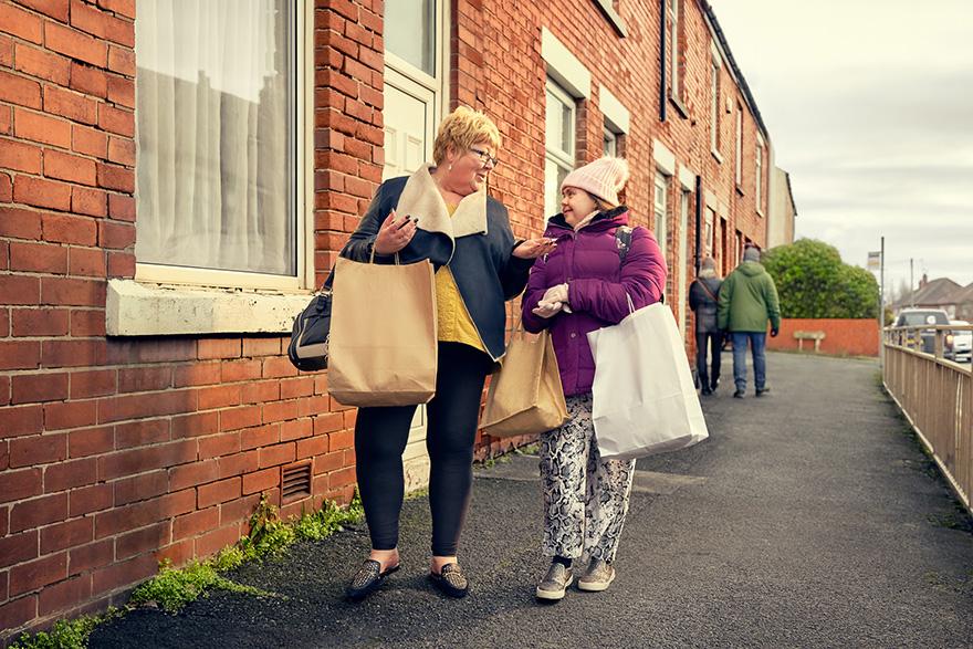 Two women, one older and one younger, standing on a street, both wearing casual clothing and holding large paper shopping bags.