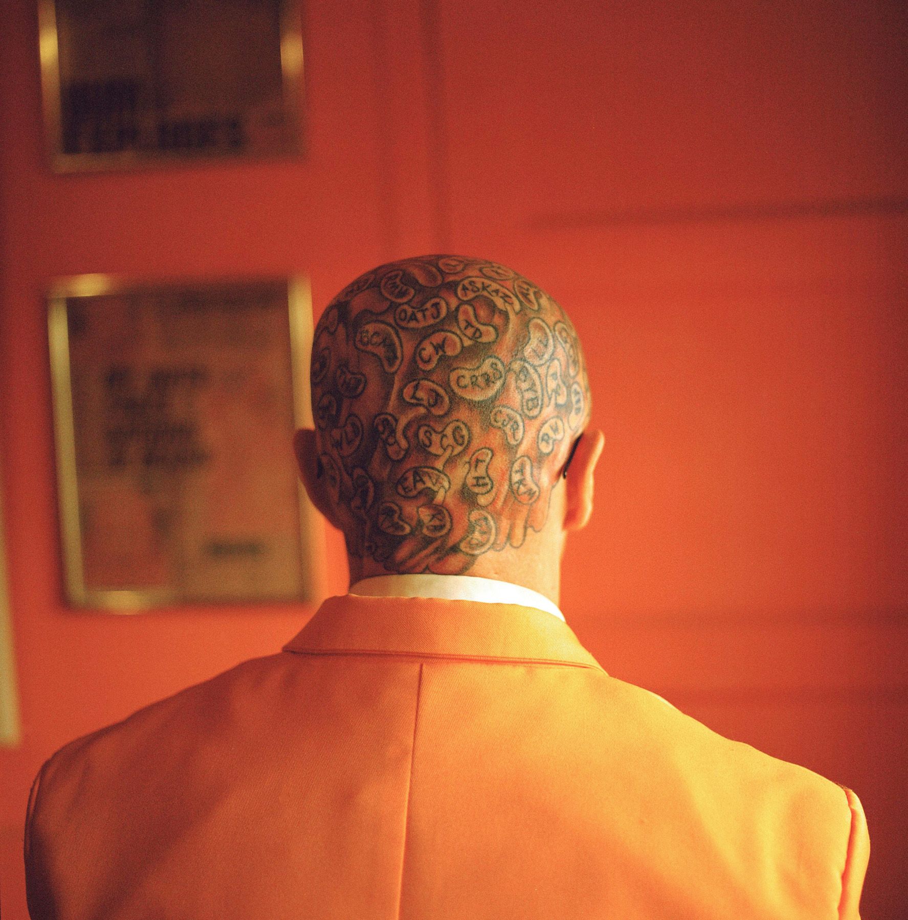 A person with tattoos stands in front of a red wall wearing a white shirt and a red jacket.
