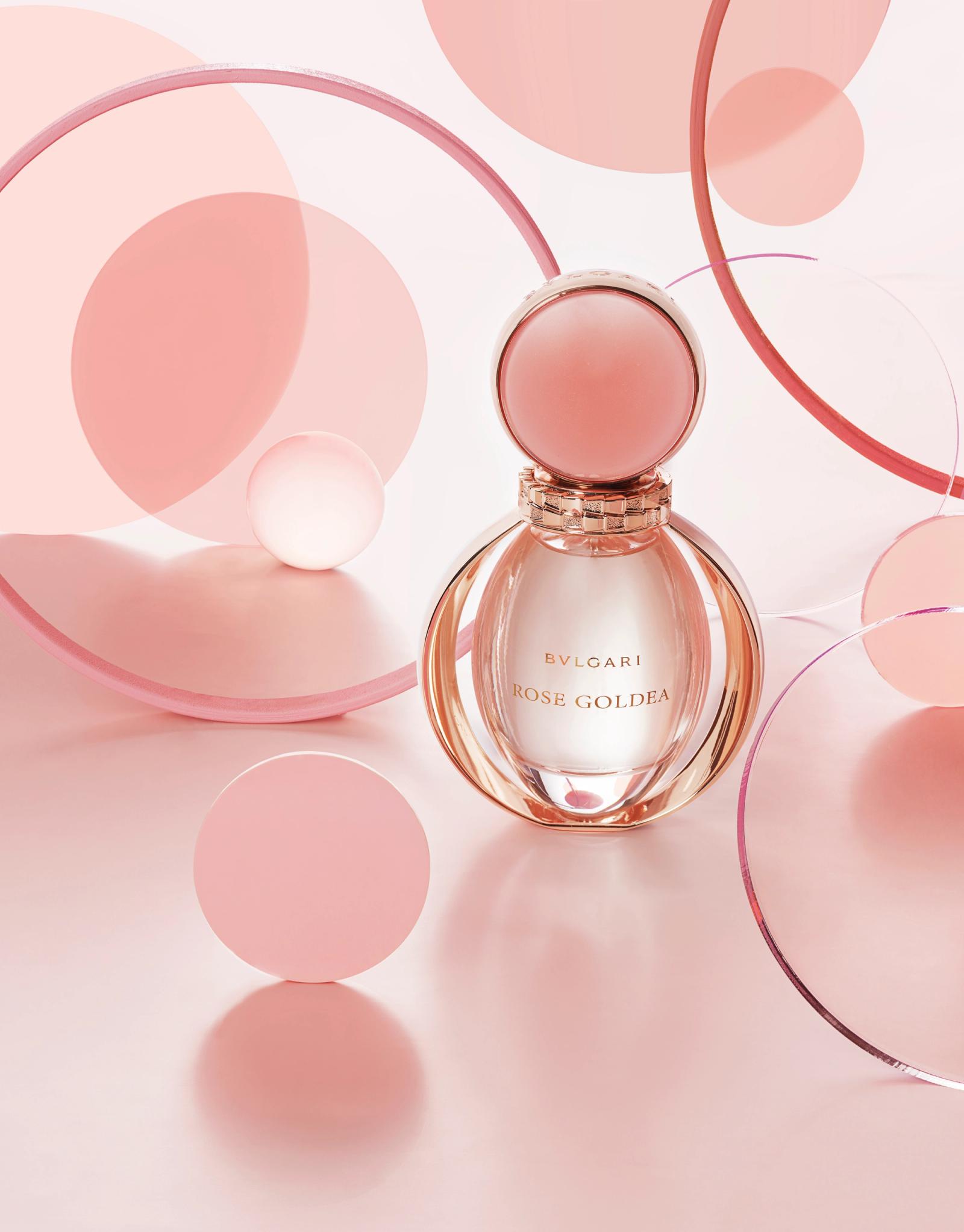 A perfume bottle with a rose gold cap and a clear glass body, surrounded by pink and white circles and a few pink and white balls.