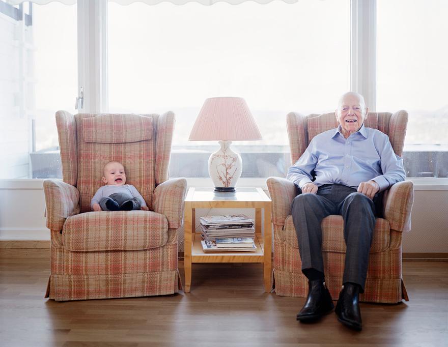 A baby in a blue onesie is sitting in a chair next to an older man in a blue shirt, both in a living room with a window in the background.