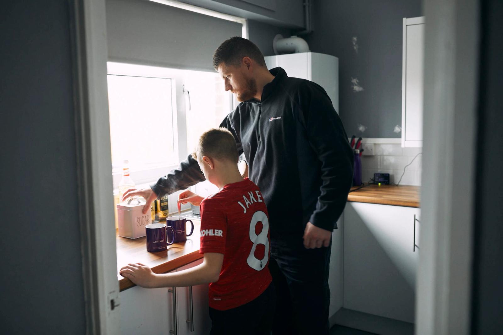 A man and a young boy stand at a kitchen counter, preparing a meal together. The boy, wearing a red t-shirt with the name "JAKE" and the number "8" on the back, looks up at the.