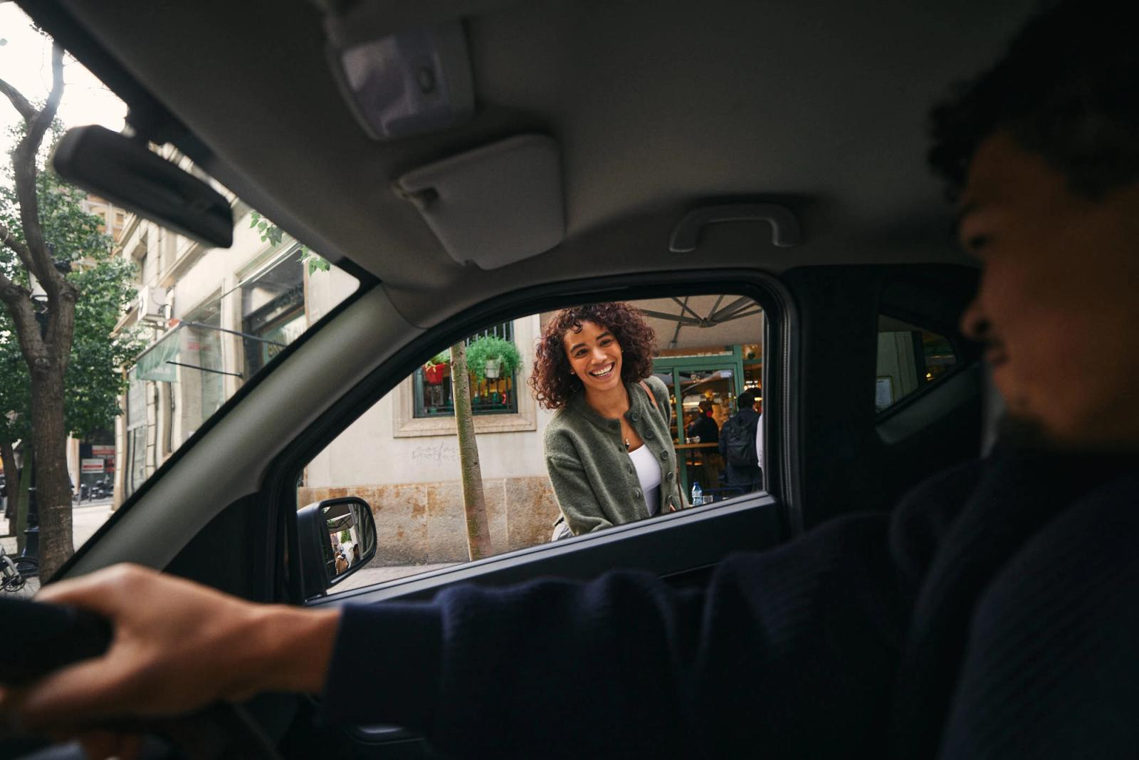 A woman with curly brown hair and a green cardigan smiles at a man through the car window. She is standing on the sidewalk, and the man is driving by, looking at her with a.