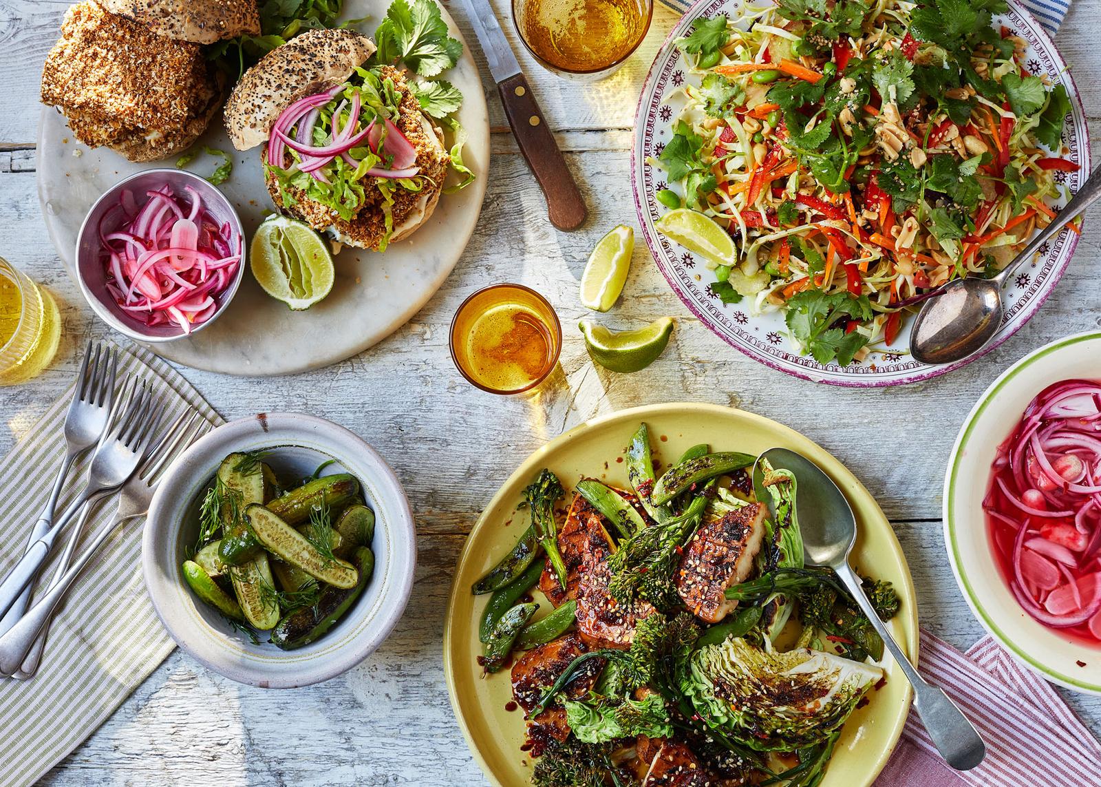 A table setting with a variety of dishes, including a burger, salad, and vegetables, on a rustic wooden table.
