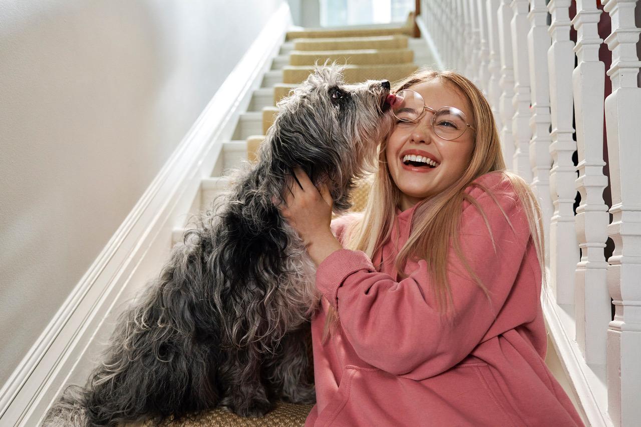 A young woman in a pink hoodie is sitting on a staircase, laughing and being licked by a small, fluffy dog.