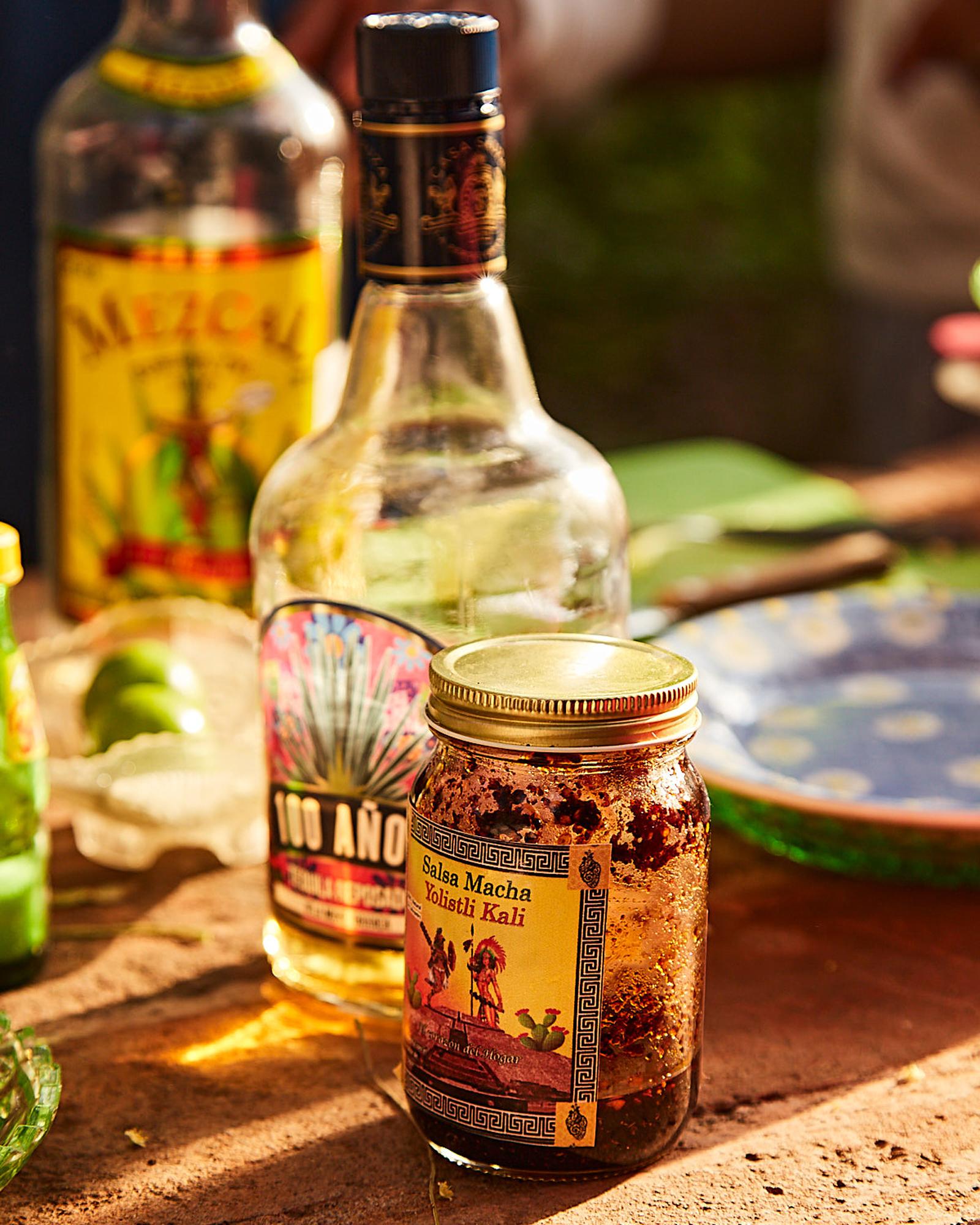 A selection of bottles of tequila and a jar of spicy sauce on a table, with a few other food and drink items in the background.