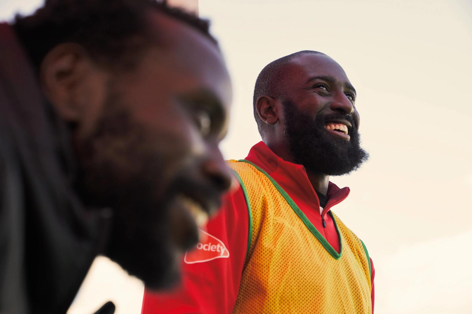 A low-angle, candid shot of two men smiling and looking upward during an outdoor session. The man in focus wears a yellow mesh training bib over a red shirt. Photograph by Nick Dolding.