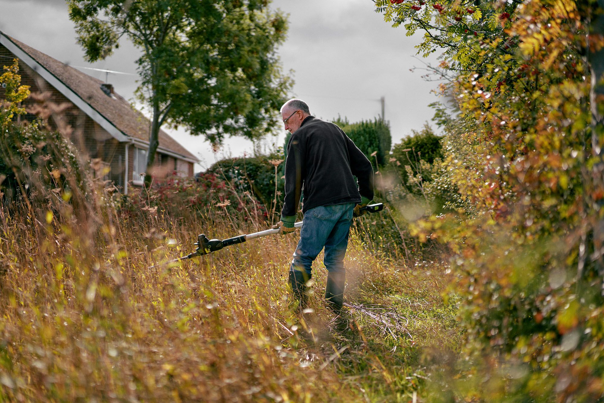 A man in a black jacket and jeans is standing in a field, holding a long, two-handed weed whacker.