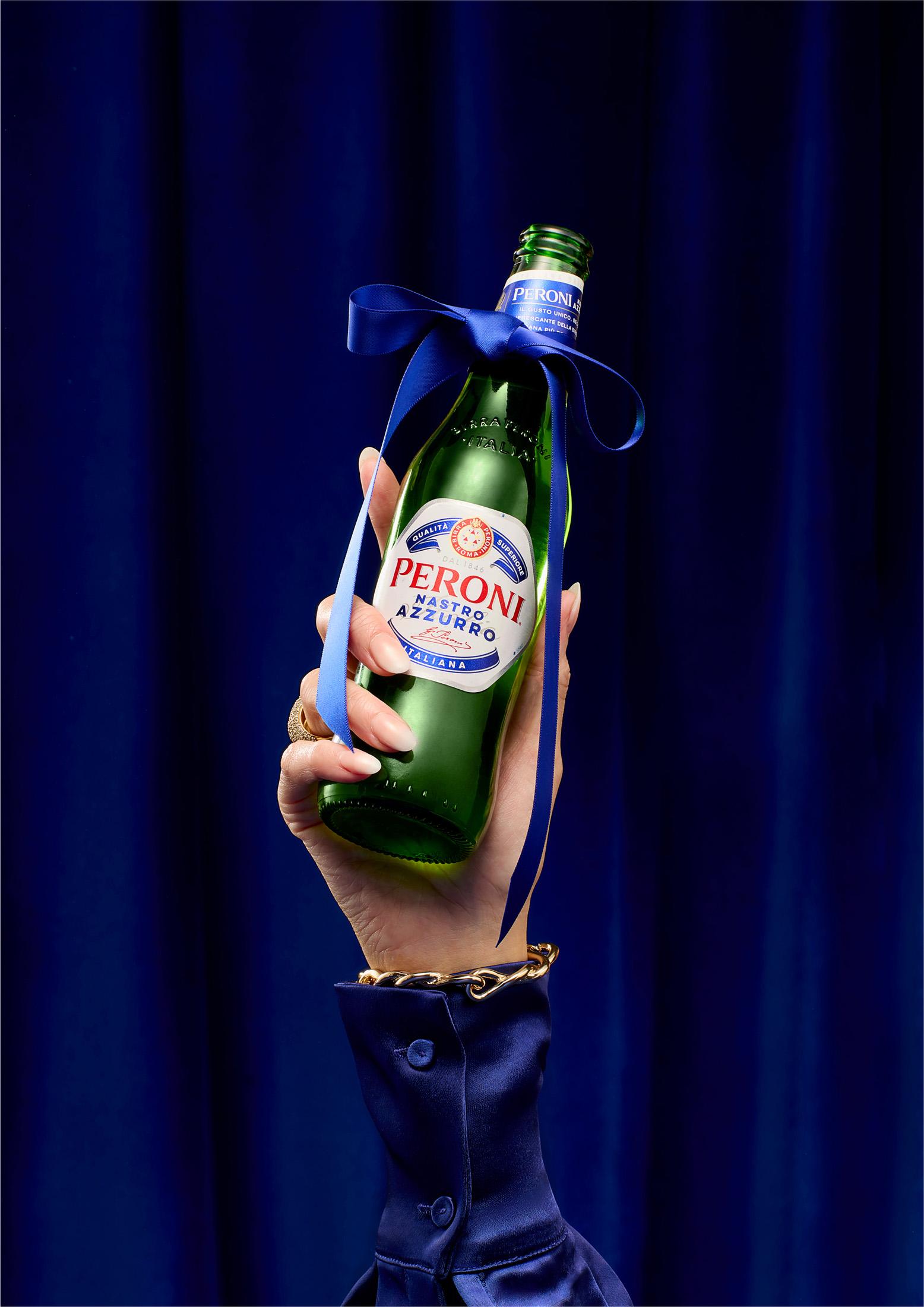 A hand holding a green glass bottle of Peroni beer, with a blue ribbon tied around the neck.