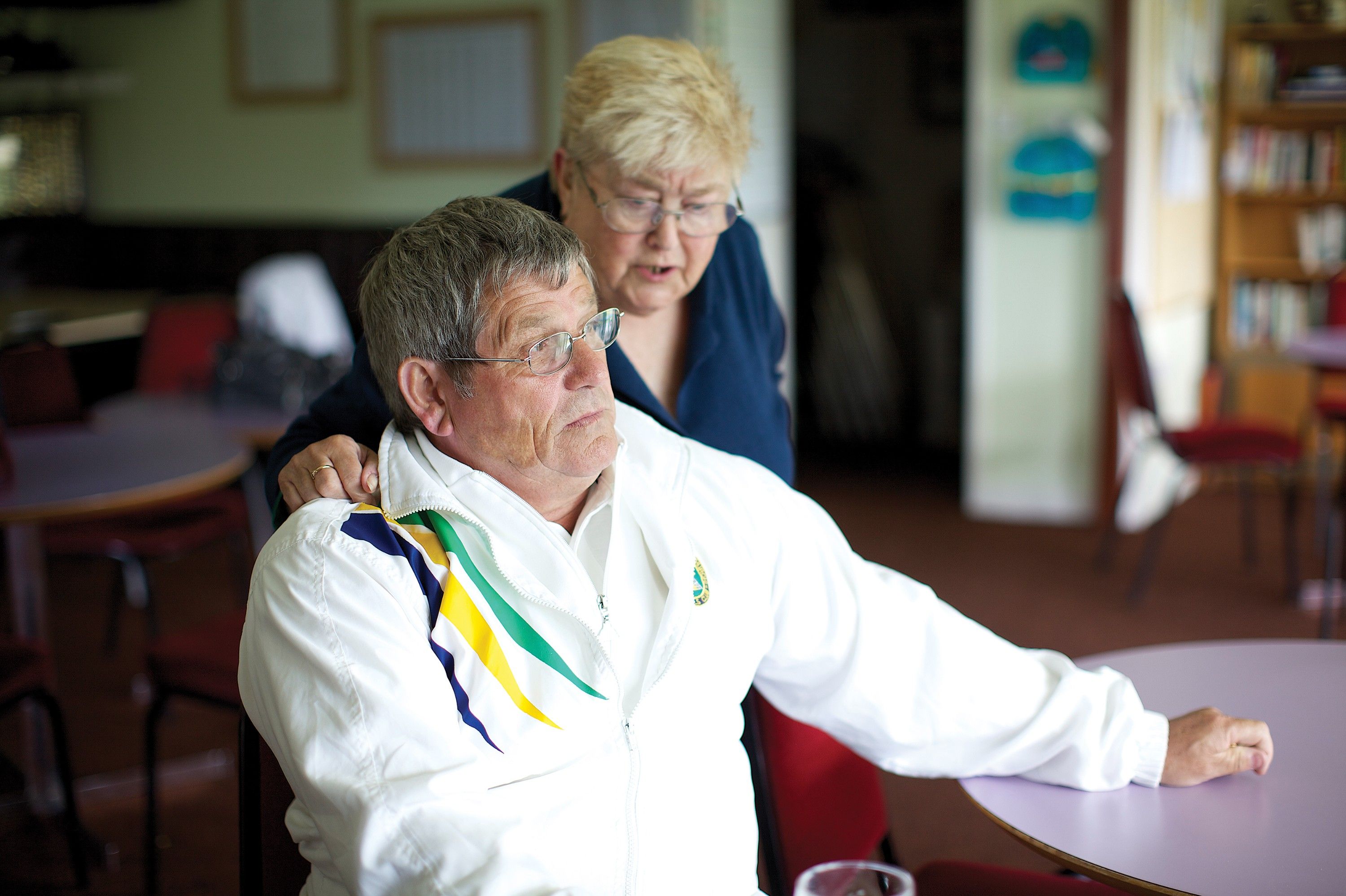 An older man in a white jacket with a multicolored stripe sits holding a woman in a blue jacket on a chair in what appears to be a room with tables and chairs.