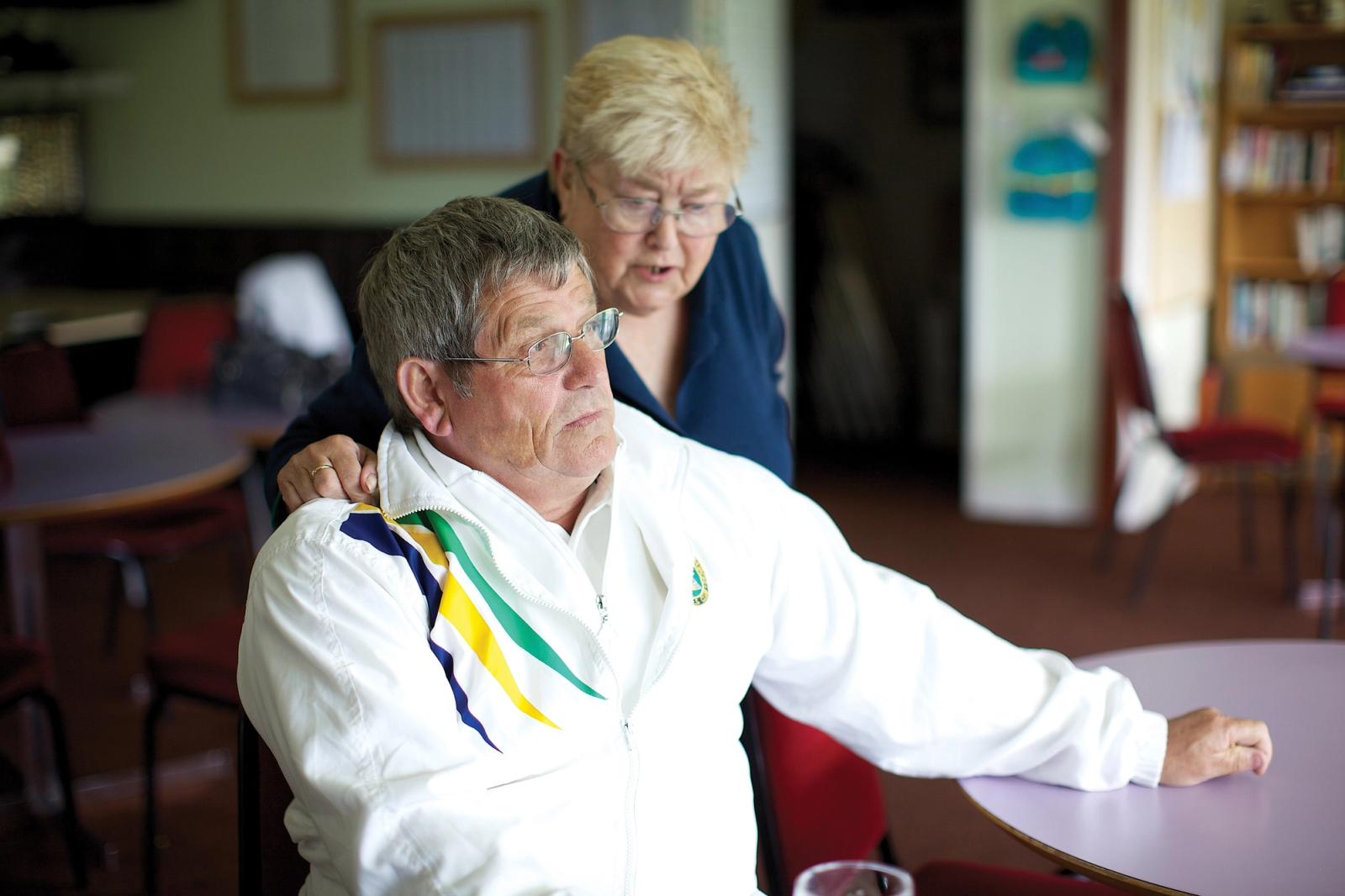 An older man in a white jacket with a multicolored stripe sits holding a woman in a blue jacket on a chair in what appears to be a room with tables and chairs.