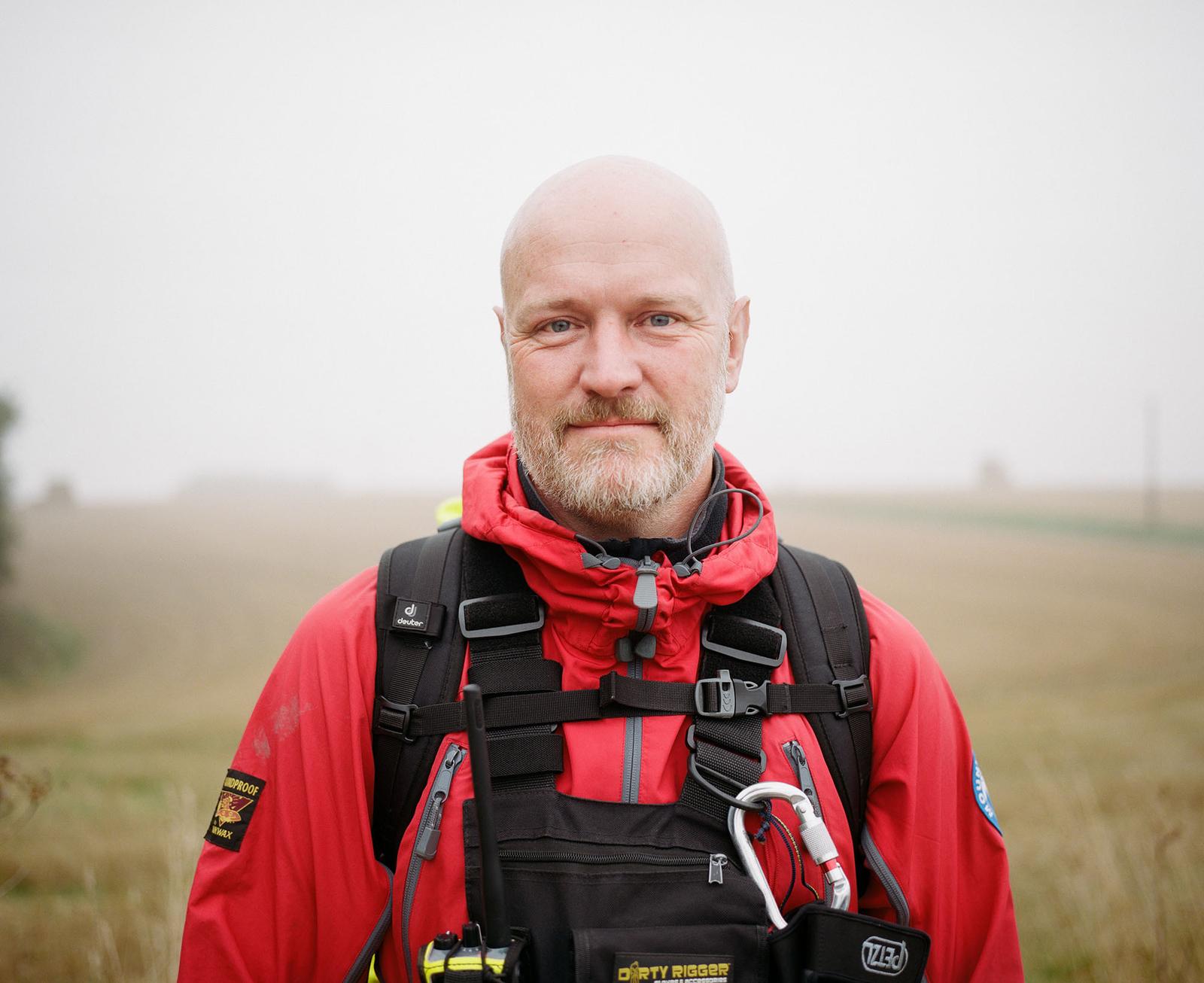 A man in a red jacket with a backpack and a camera around his neck, standing in a field of tall grass.