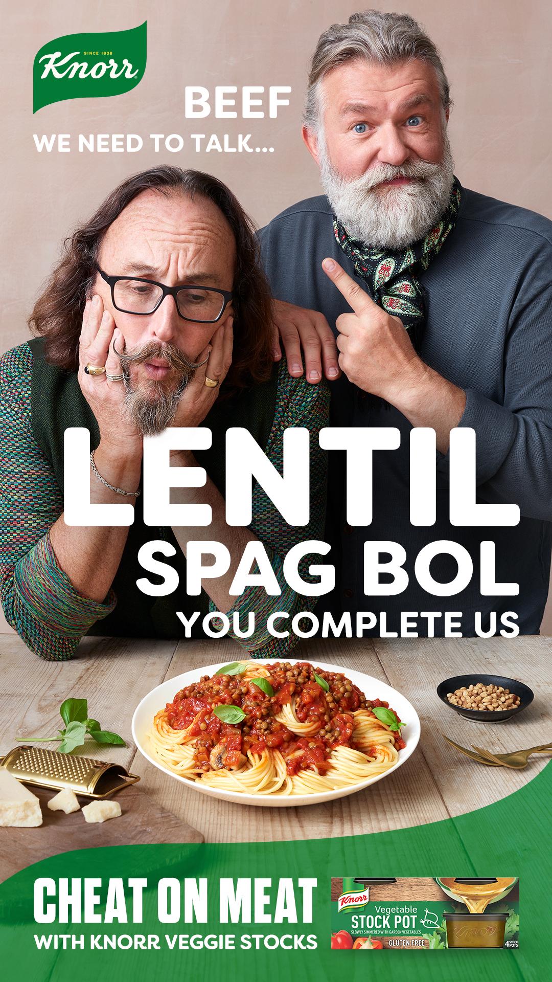 Two men, one with a beard, sitting at a table with a plate of spaghetti and a bowl of lentil spagbol, with a background of a beige wall.