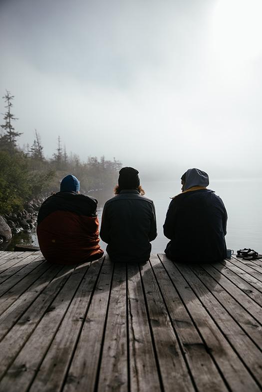 Three people sitting on a dock, looking out at a foggy lake.