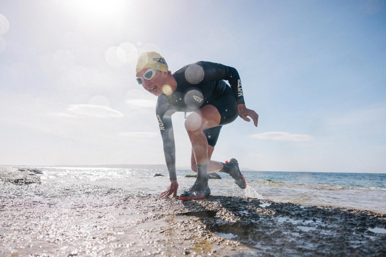 A man in a wetsuit and swim cap is running on a rocky beach, preparing to enter the water for a swim or run in the ocean.