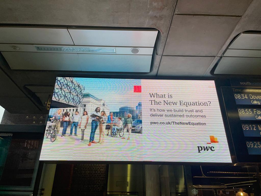 A large digital screen in a train station displaying a PWC advertisement with a diverse group of people in a city setting, with a city skyline in the background.