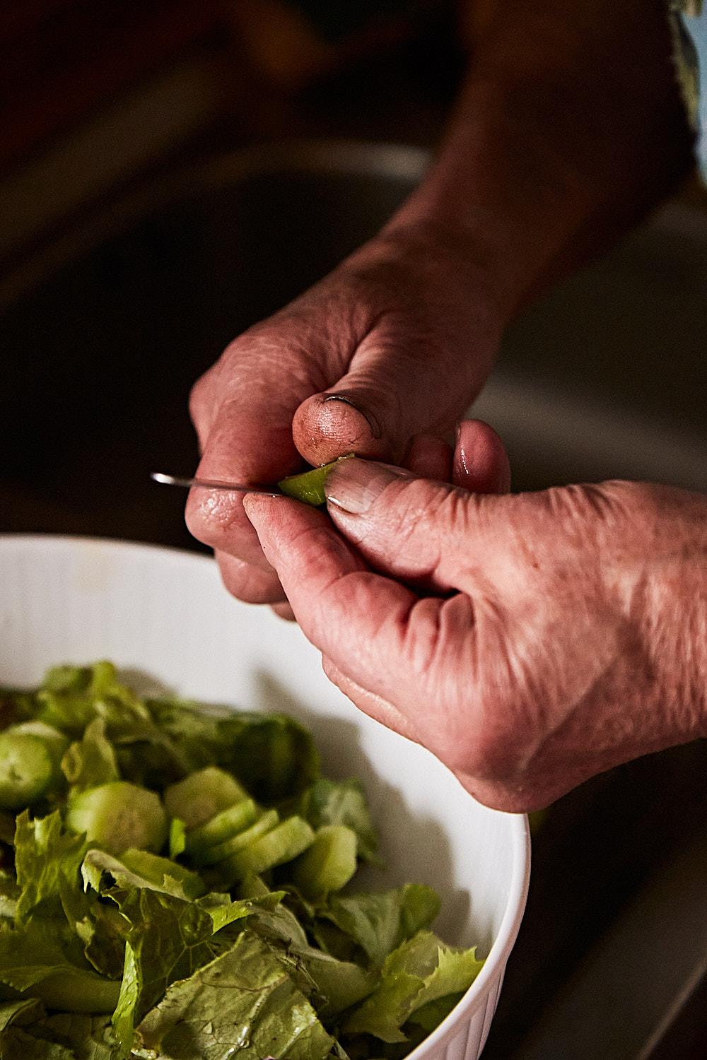 A person's hands are carefully peeling a green leaf from a lettuce head, with a white bowl of lettuce in the background.