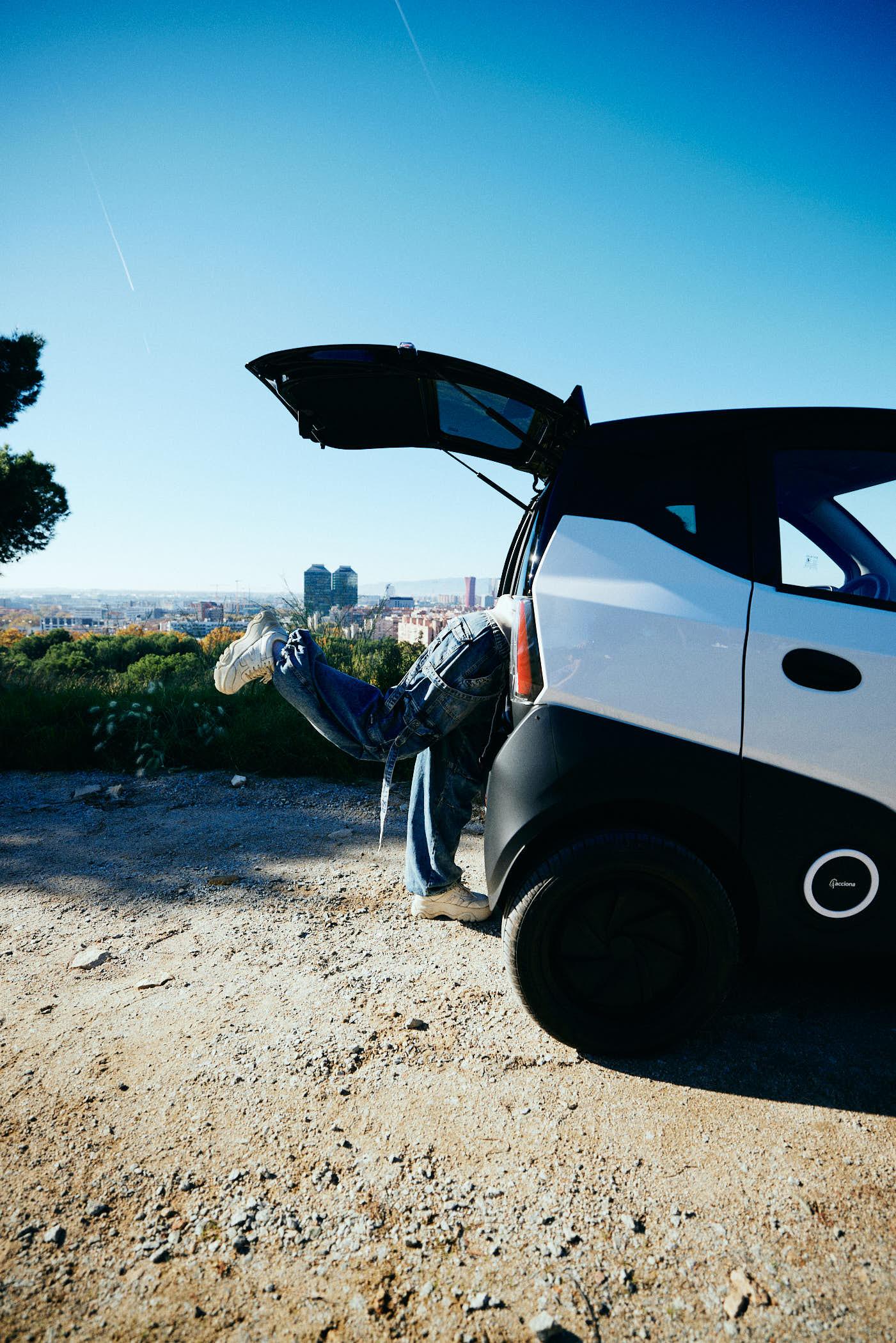 A person is leaning out of the open trunk of a white car, with their legs and feet visible. They are wearing blue jeans and white sneakers, and are standing on a dirt road with a.