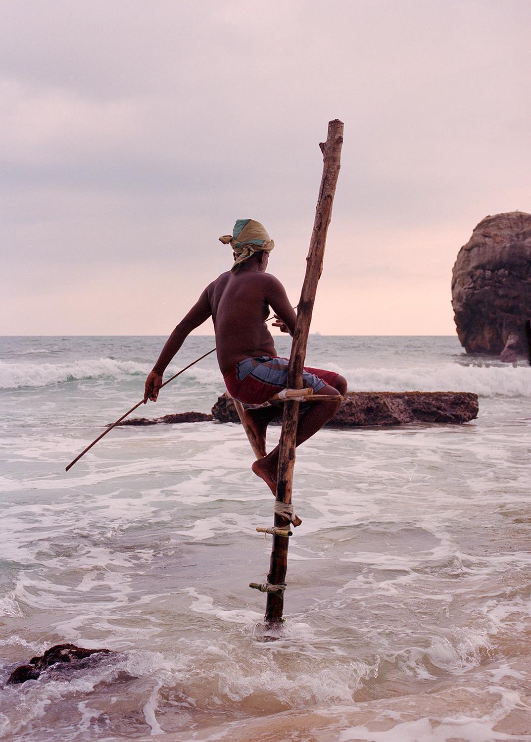 A man in a red and blue striped sarong and a turban-like headscarf is perched on a wooden pole in the ocean, using a long pole to catch fish.