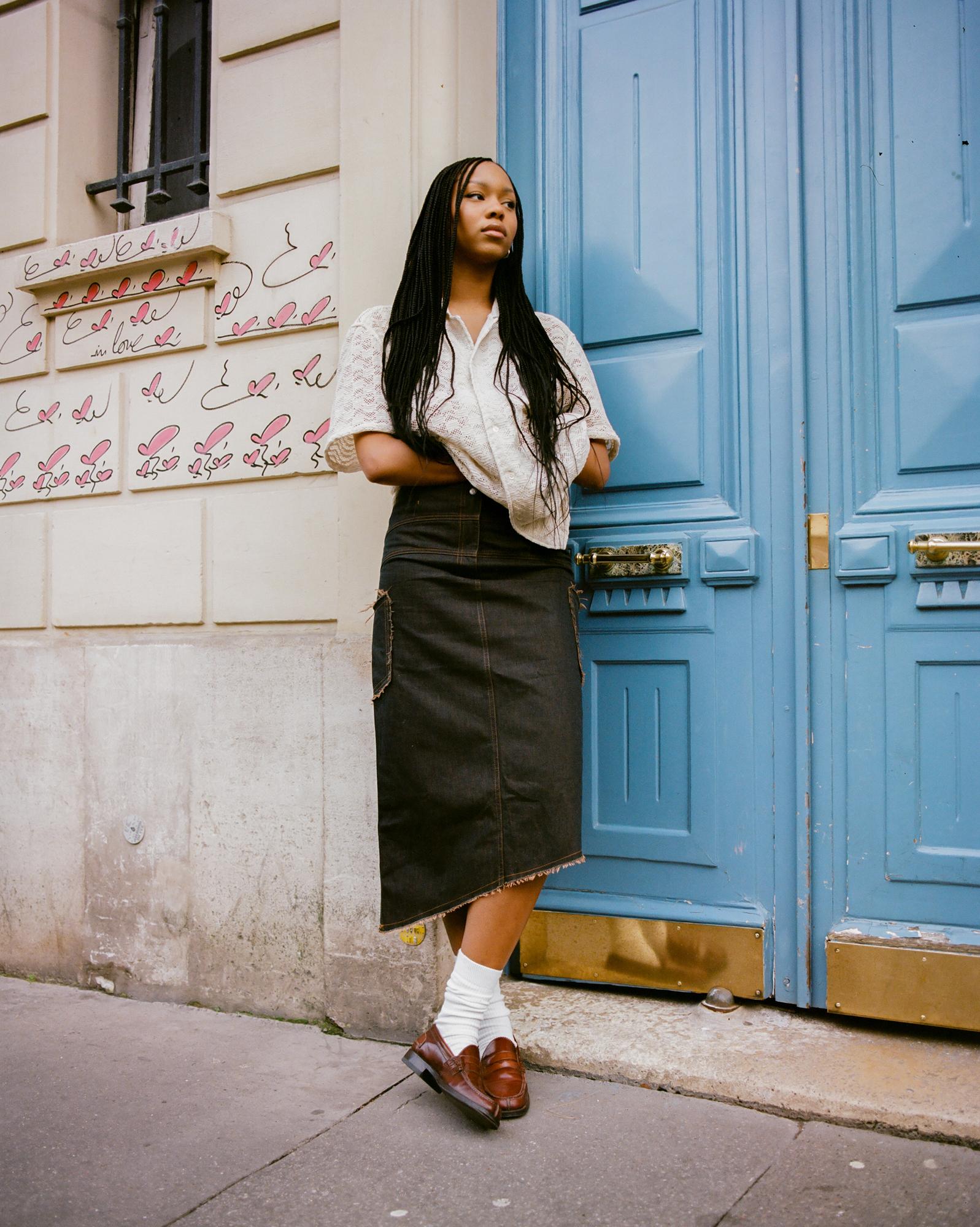 A woman stands in front of a blue door wearing a white lace top, black denim skirt, and brown loafers.