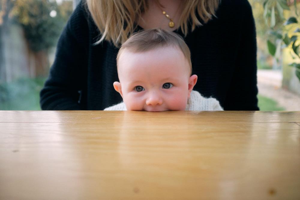 A baby peeks over a wooden table, looking up at the camera with wide, curious eyes. A woman with blonde hair and a black sweater stands behind the table, out of focus.