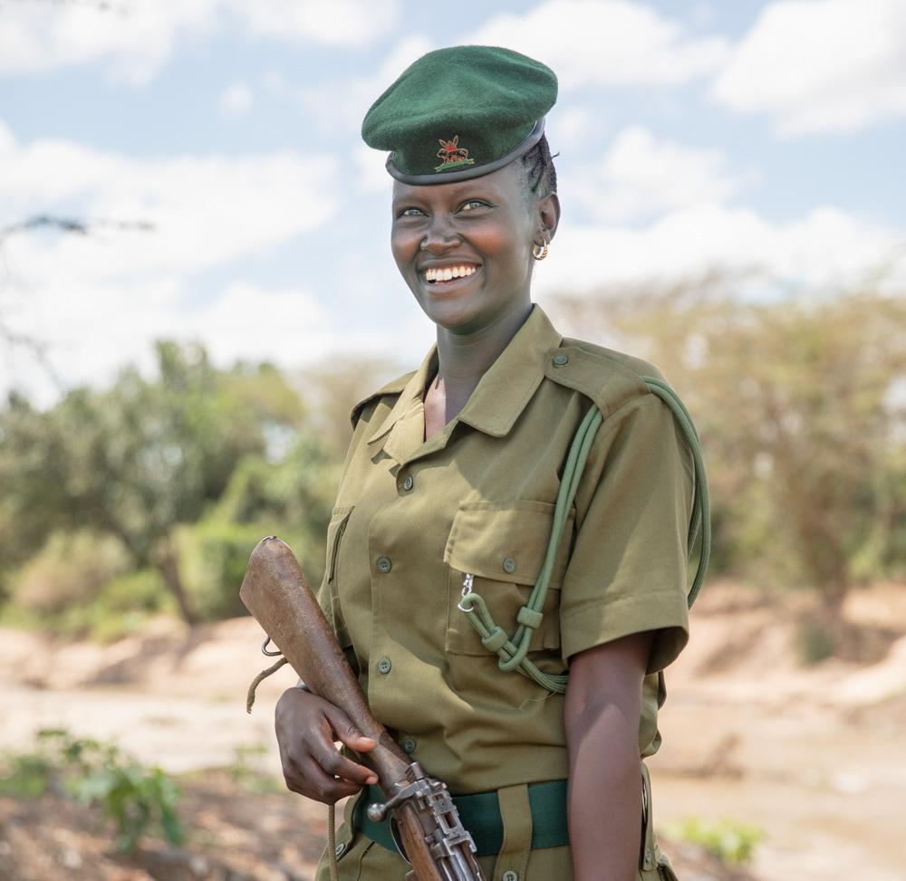 A woman in a military uniform, holding a rifle, in a desert or savannah-like environment.