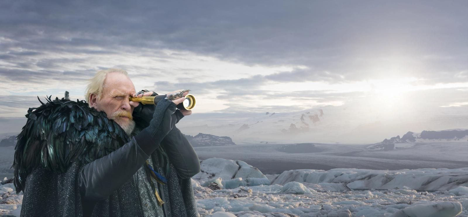 A man wearing a black cloak and pointed hat examining something with a similar cylindrical object, in a snowy mountain setting.
