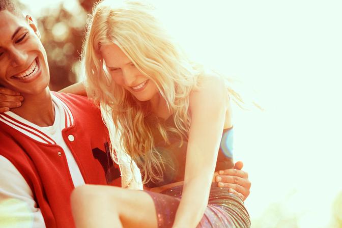 A young woman with long blonde hair is being lifted by a young man with a football jersey, both smiling and laughing.