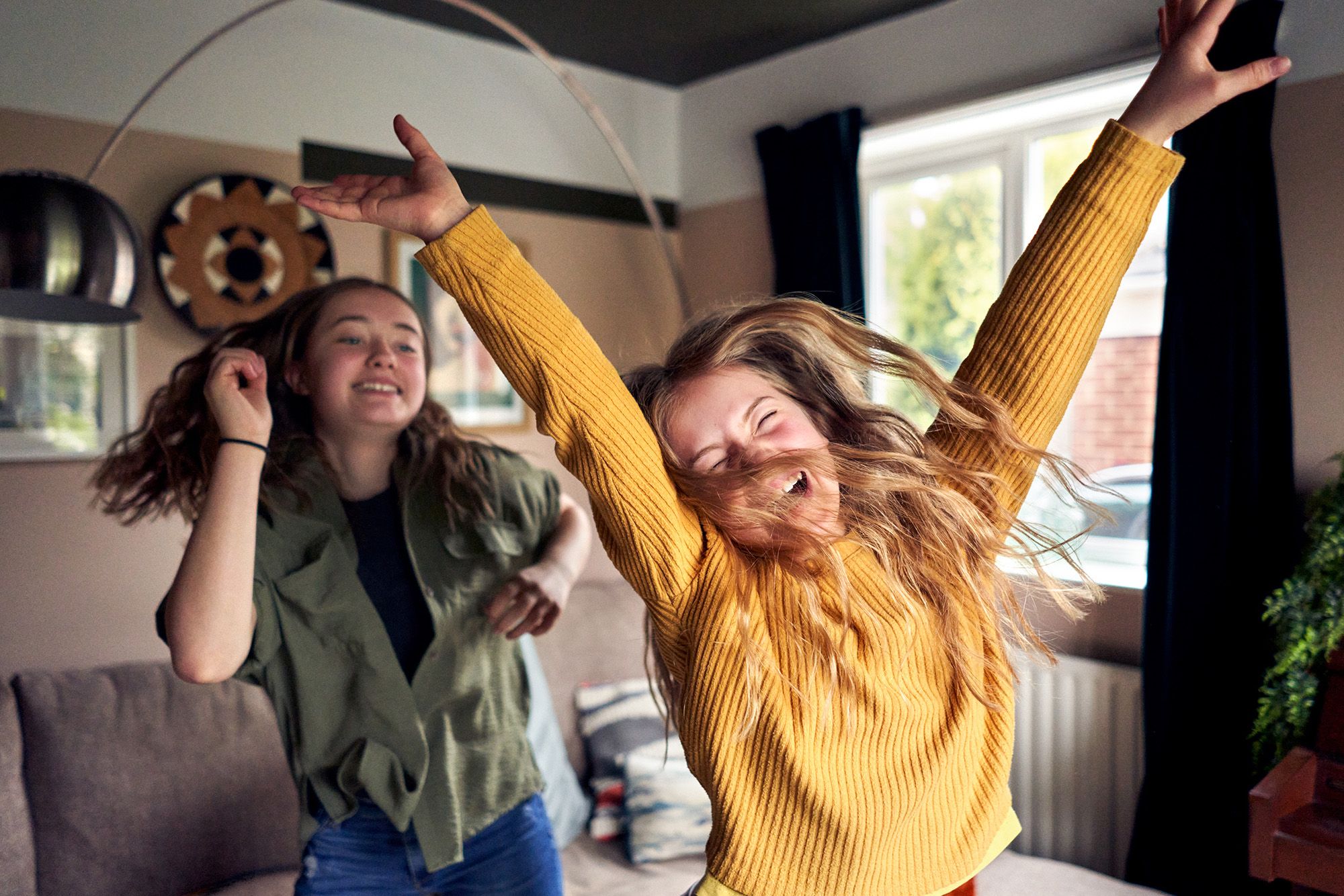 Two women dance and laugh in a living room, one wearing a yellow sweater and the other a green jacket.