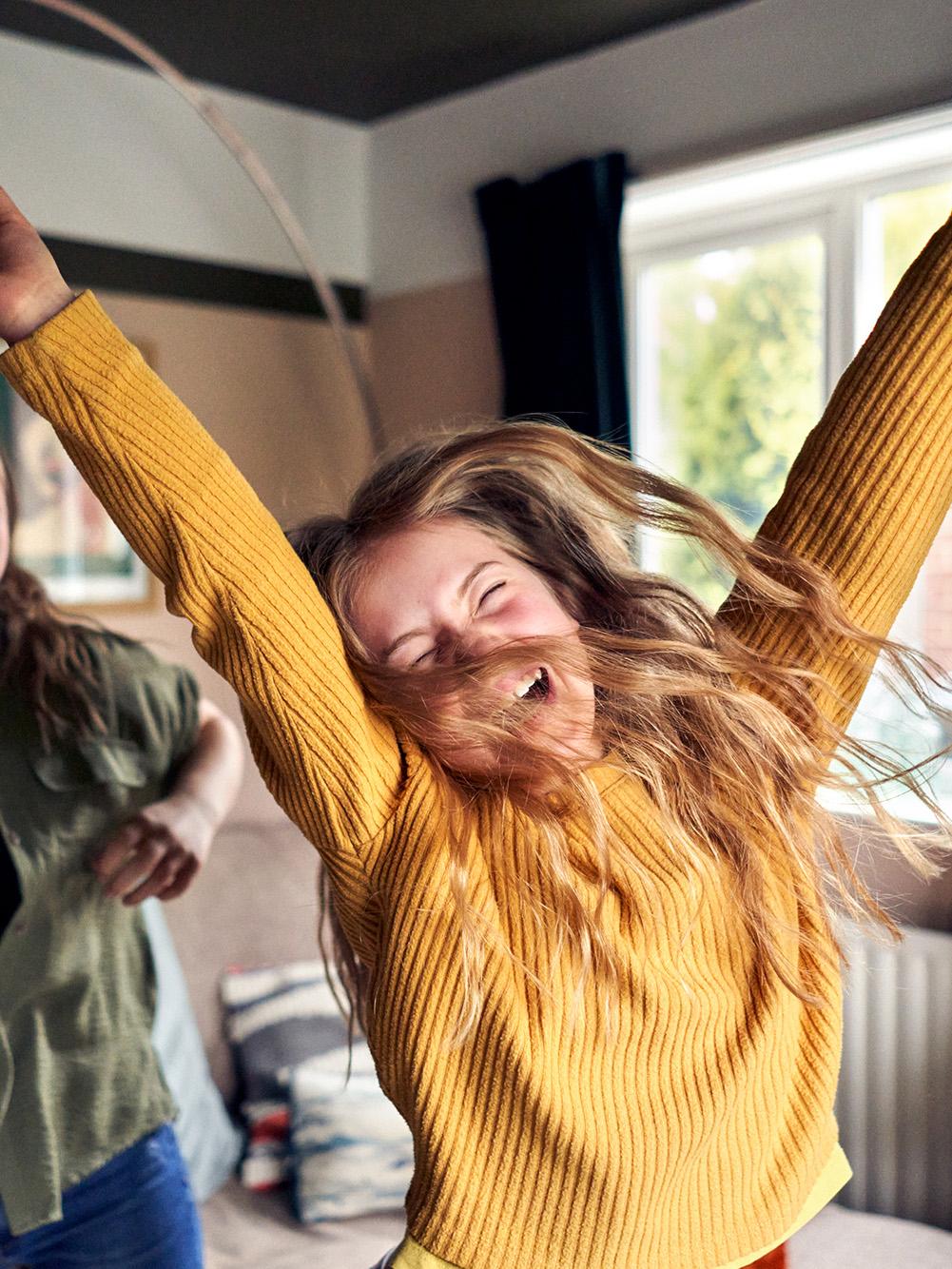 Two women dance and laugh in a living room, one wearing a yellow sweater and the other a green jacket.