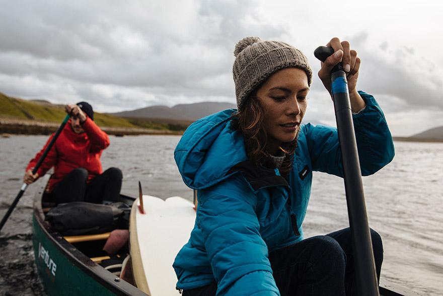 Two people in a canoe on a lake, one woman in a blue jacket and grey beanie, the other in a red jacket and black hat, both holding paddles.