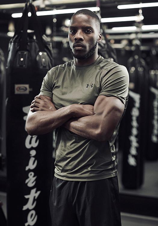 A man in a gym, wearing a grey t-shirt and black shorts, with his arms crossed and a serious expression.