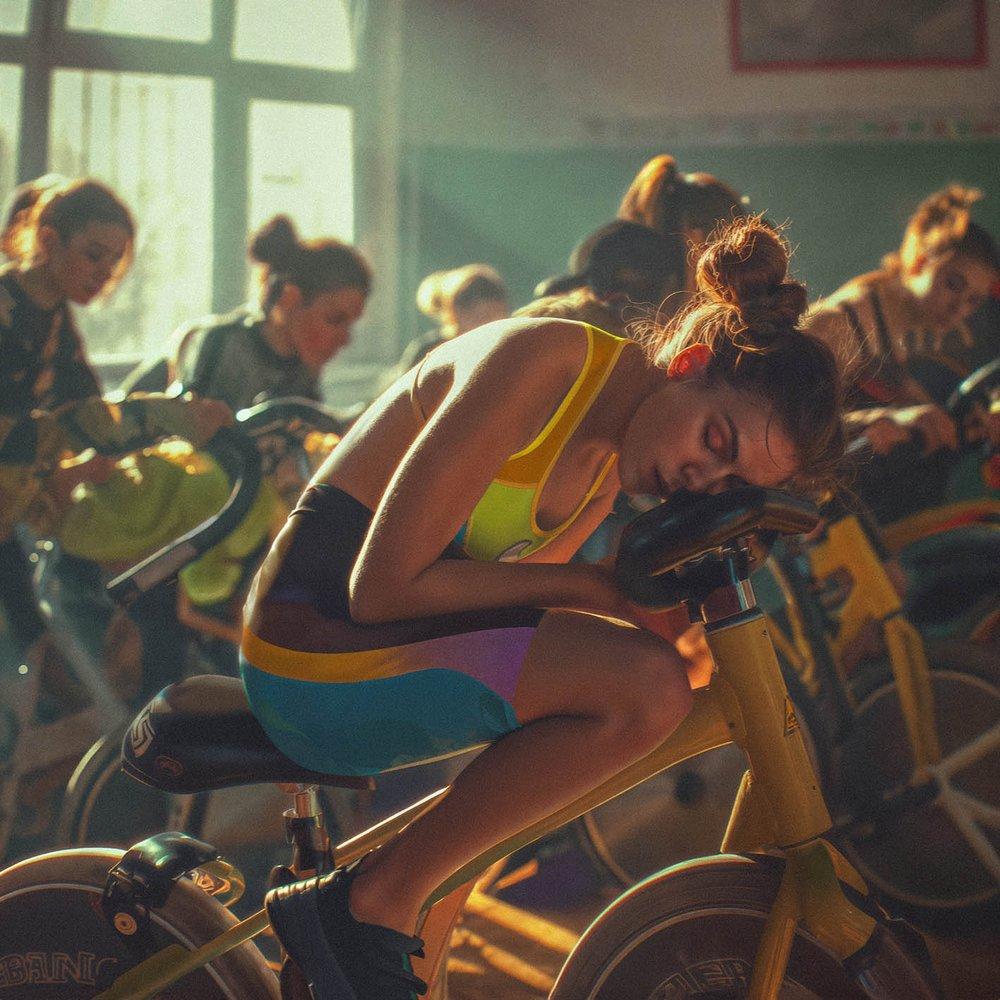 A woman in a bright yellow and blue sports bra and matching shorts is slumped over a stationary bike, exhausted.