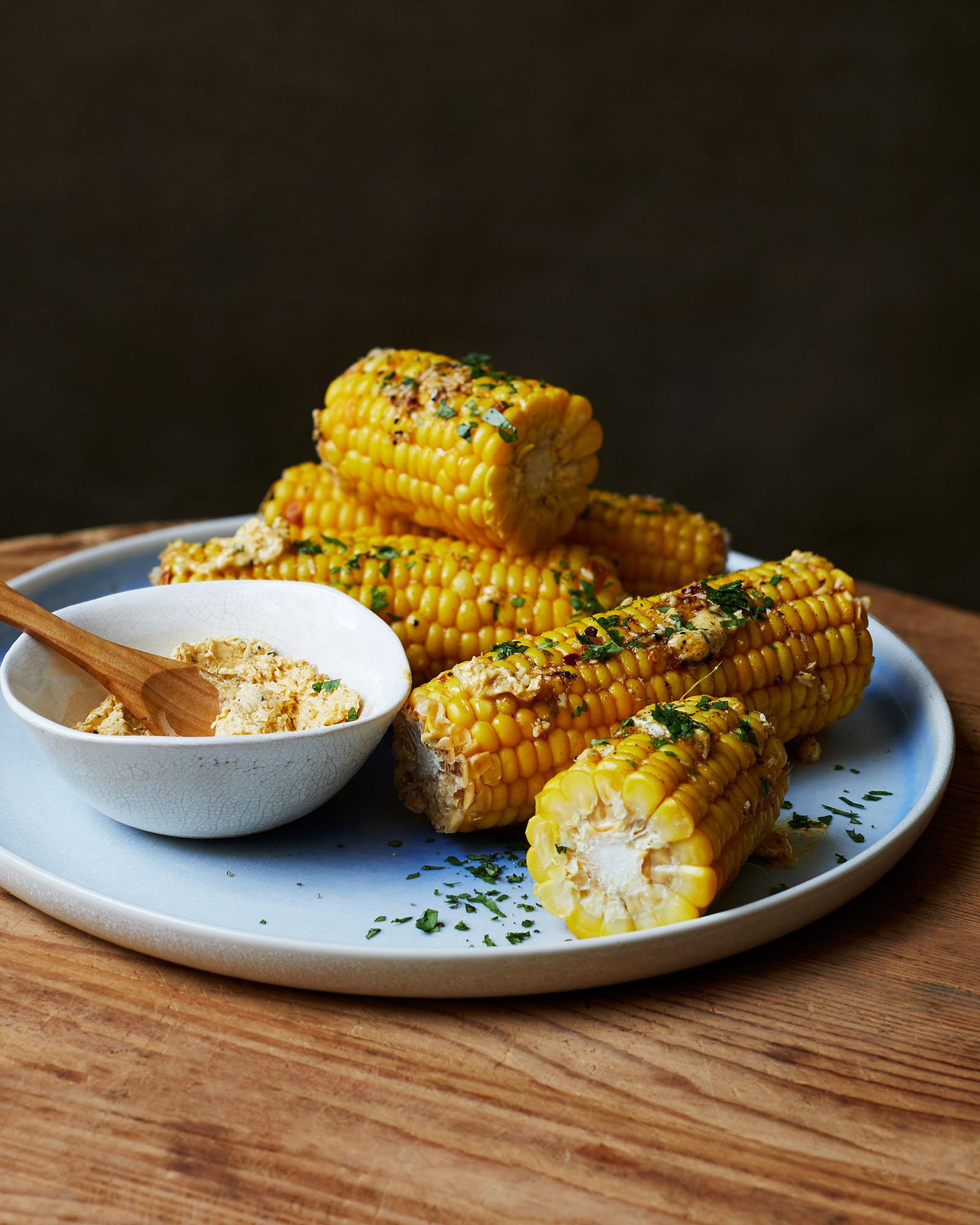 A plate of roasted corn on the cob, topped with a creamy, crumbly, and spicy sauce, served with a side of butter and a few fresh herbs.