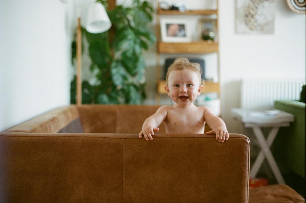 A happy, shirtless baby standing in a brown couch, looking directly at the camera with a big smile.
