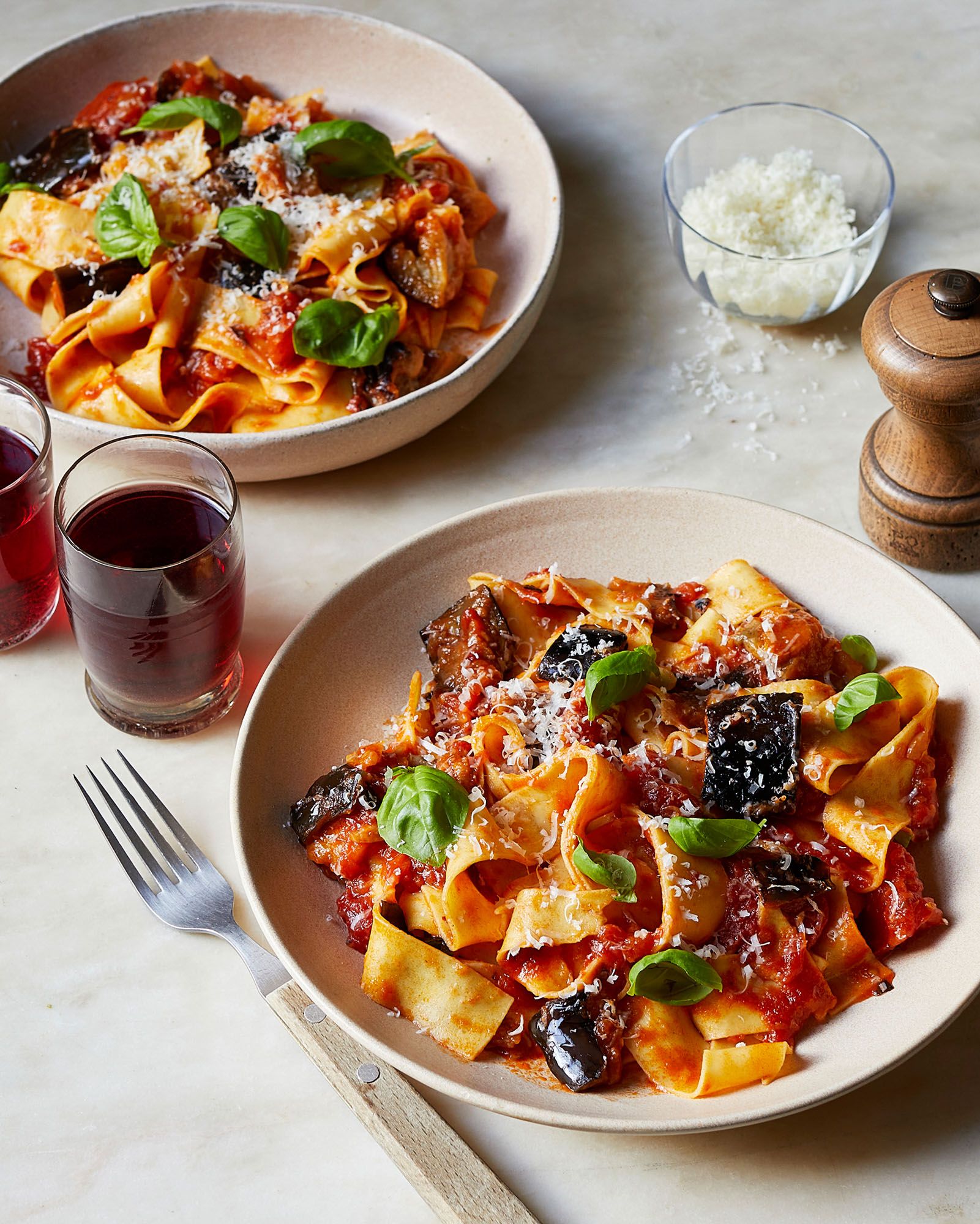 A plate of pasta with a red sauce, topped with parmesan cheese and basil leaves, served on a white table with a glass of red wine and a fork.
