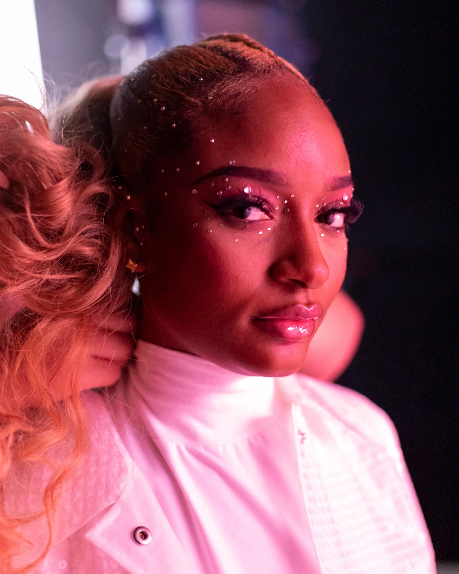 A woman with a shaved head and a white shirt, with a woman in the background with long hair, both with makeup and glitter on their faces.