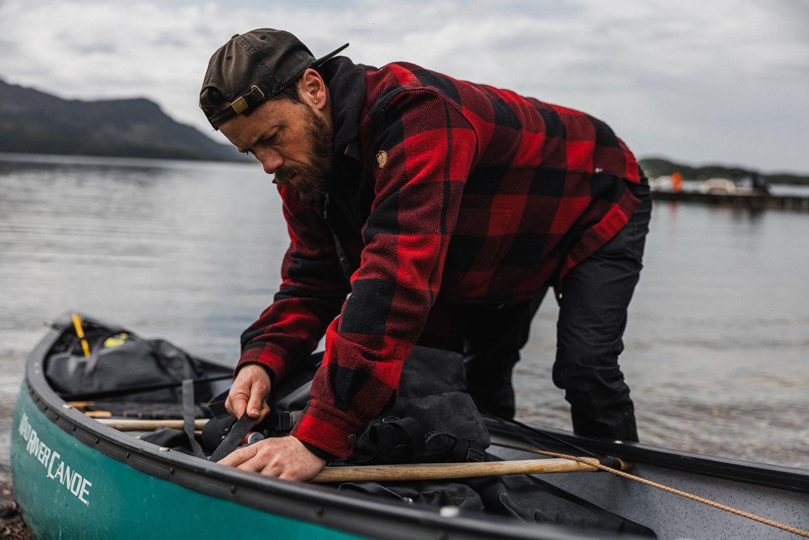 A man in a red and black plaid shirt and a black baseball cap is preparing a canoe on a lake shore, with a mountain in the background.