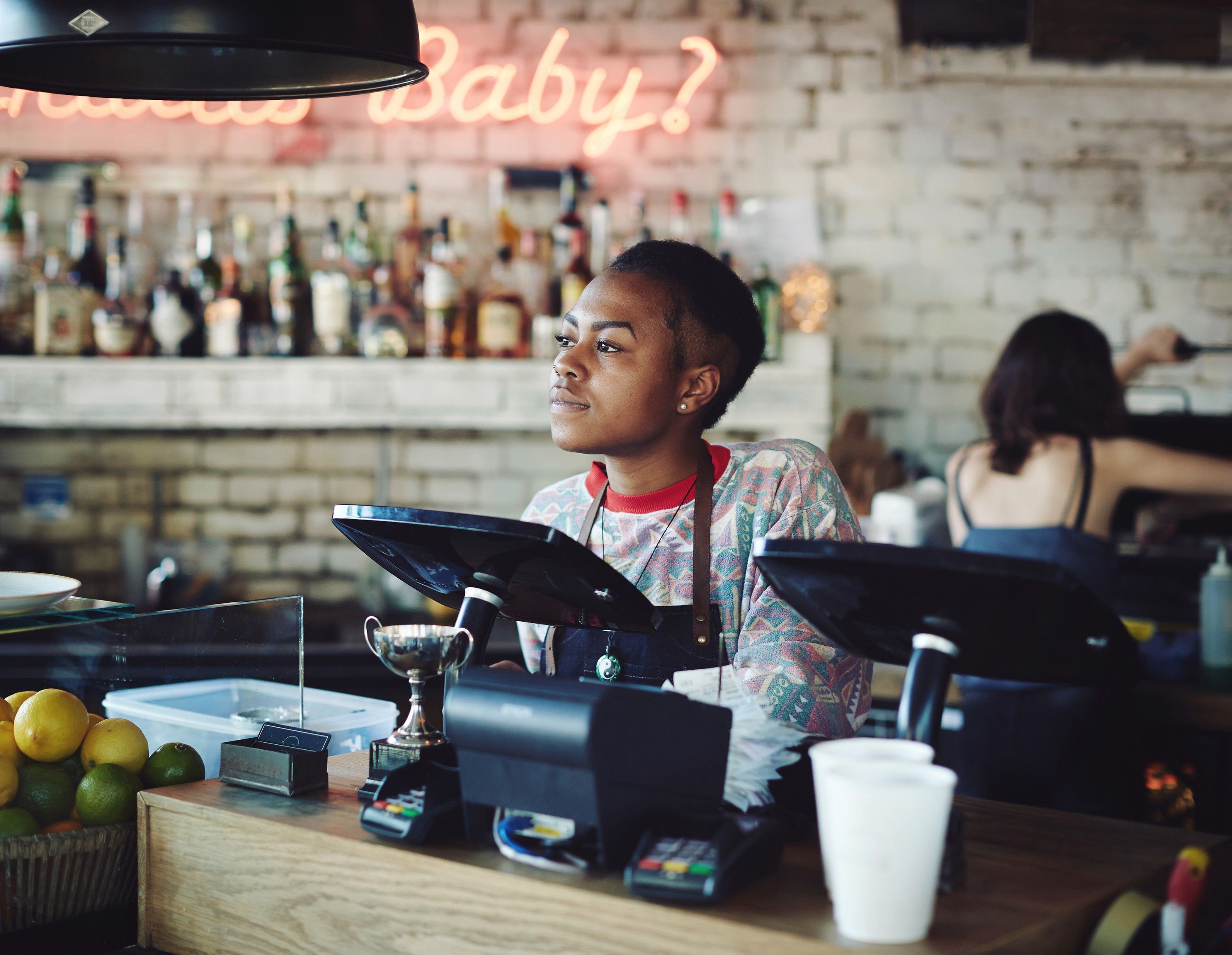 A woman with short hair and a colorful, patterned shirt is working behind a bar, surrounded by bottles of liquor and a neon sign reading 'Baby?' in the background.