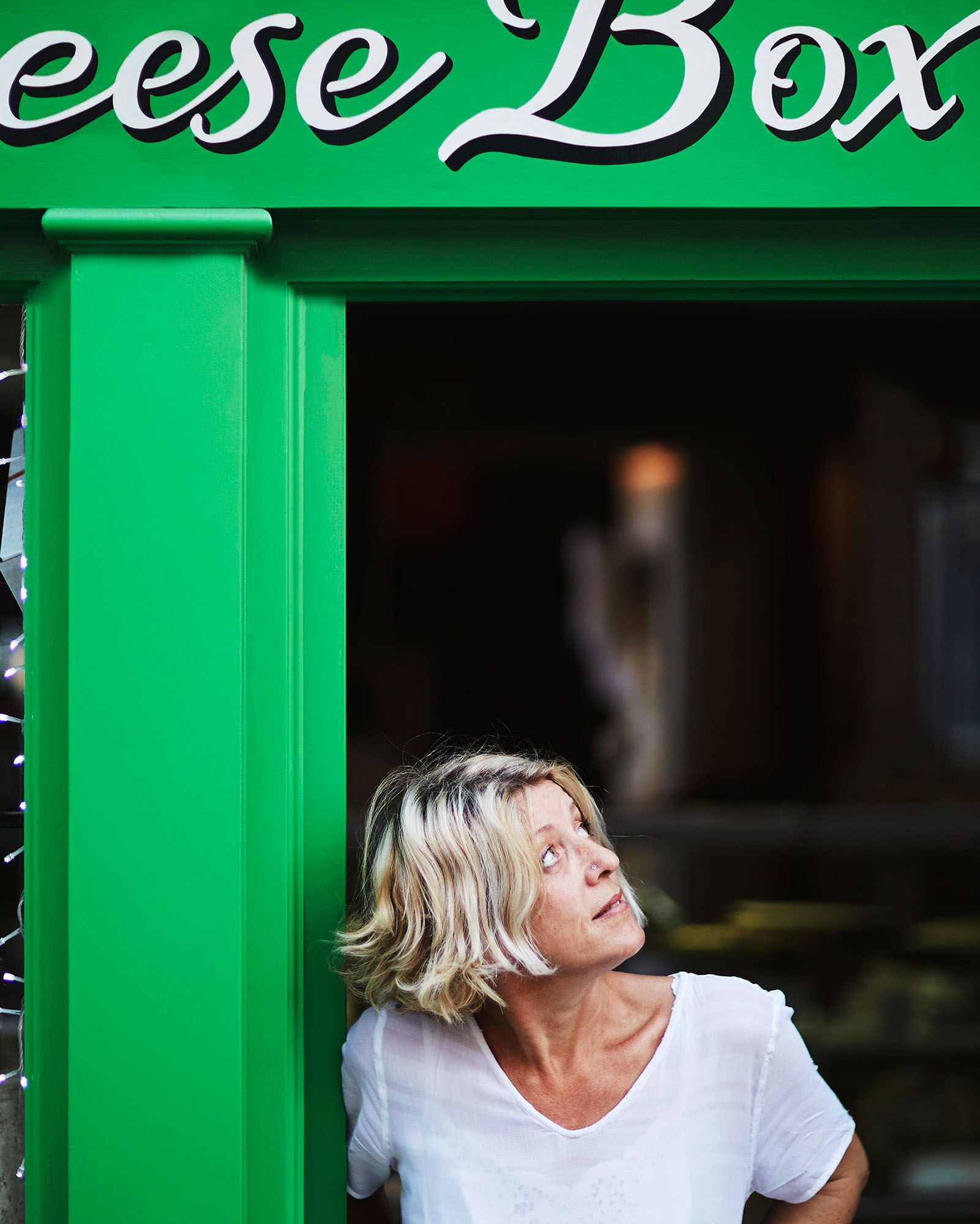 A woman stands in front of a green door with a sign that reads 'Cheese Box'.
