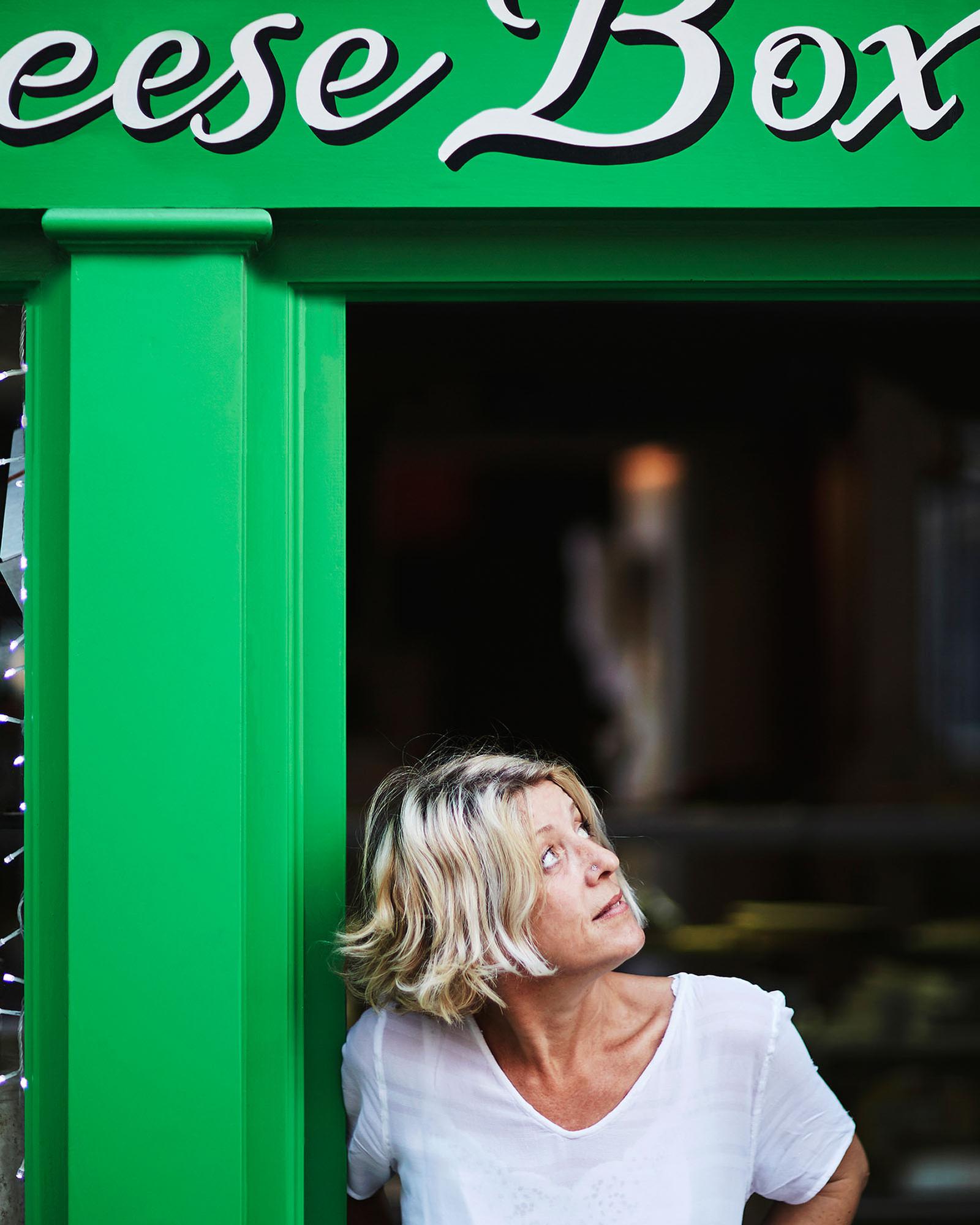 A woman stands in front of a green door with a sign that reads 'Cheese Box'.