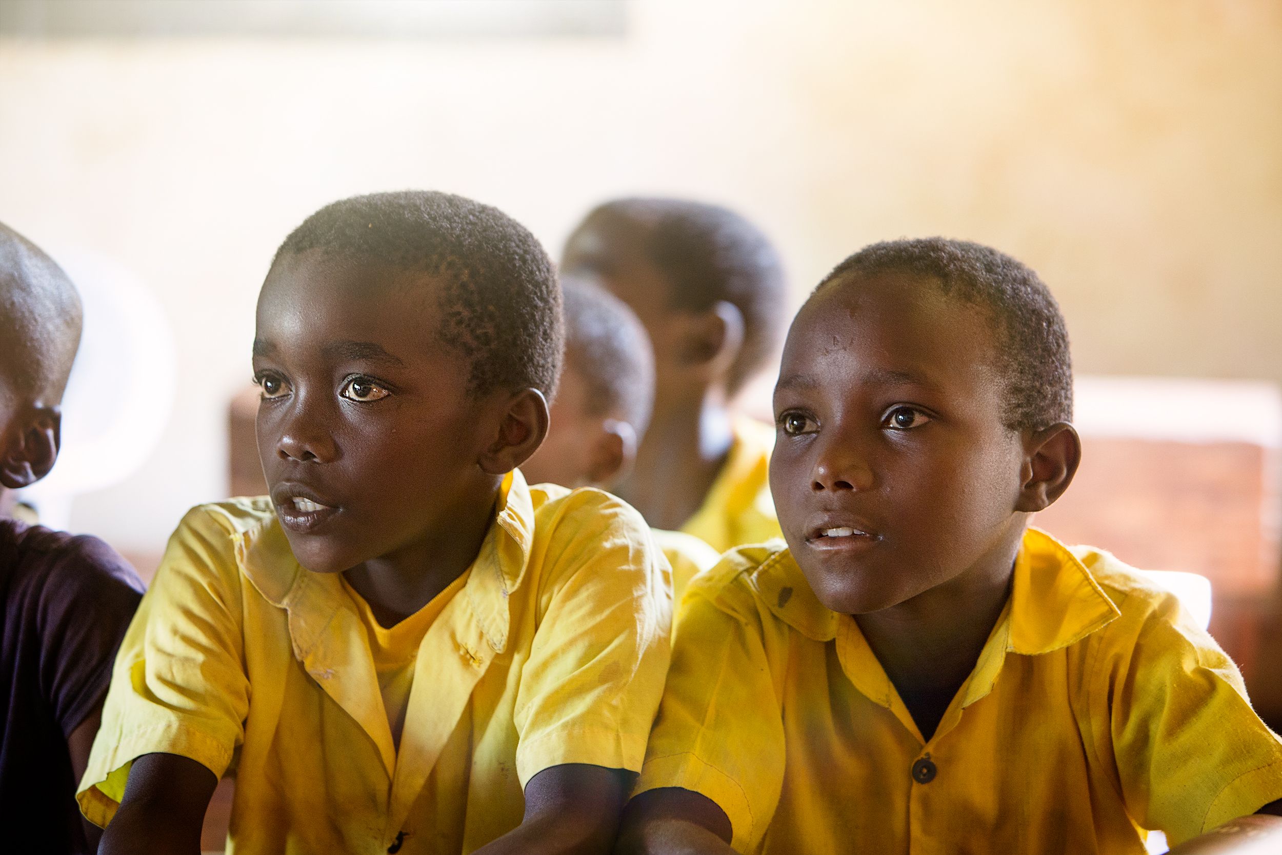 A group of young, black boys in yellow school uniforms are sitting in a classroom, looking at something off-camera with a mix of curiosity and concern.