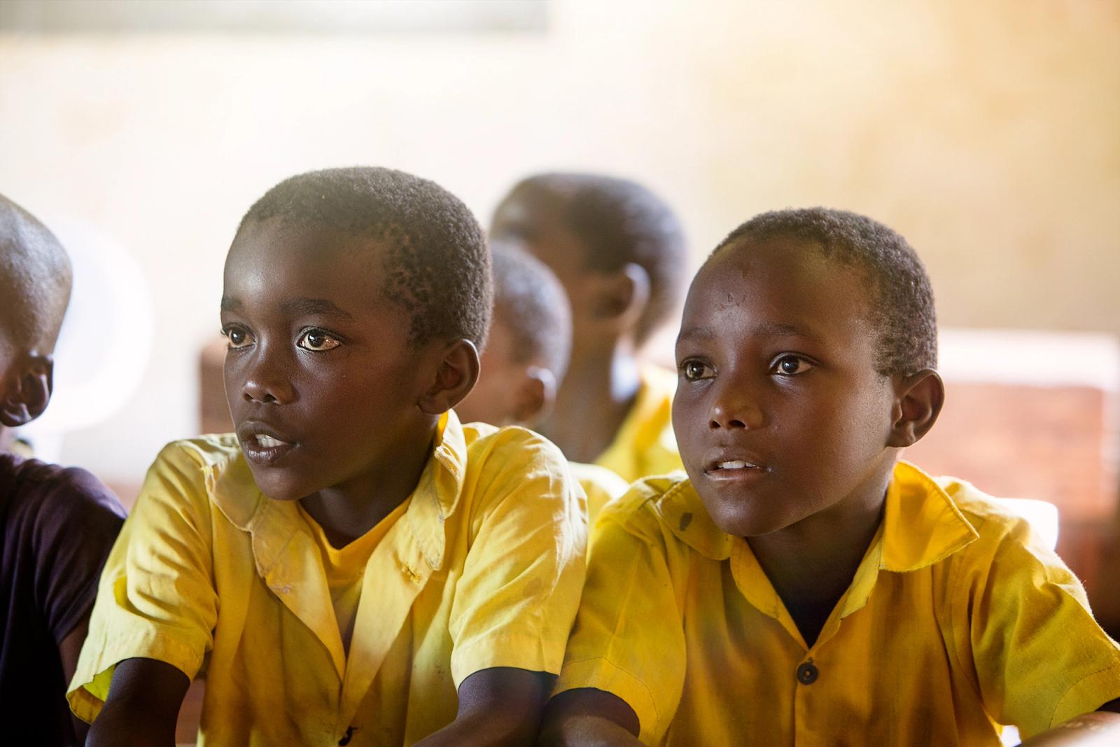 A group of young, black boys in yellow school uniforms are sitting in a classroom, looking at something off-camera with a mix of curiosity and concern.