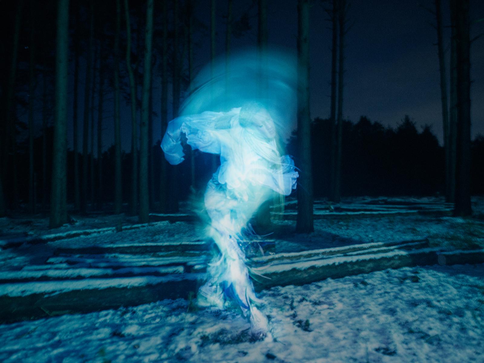 A ghostly figure in a forest, wearing a long, flowing white gown, with a hood and a cloak, surrounded by trees and a misty atmosphere.