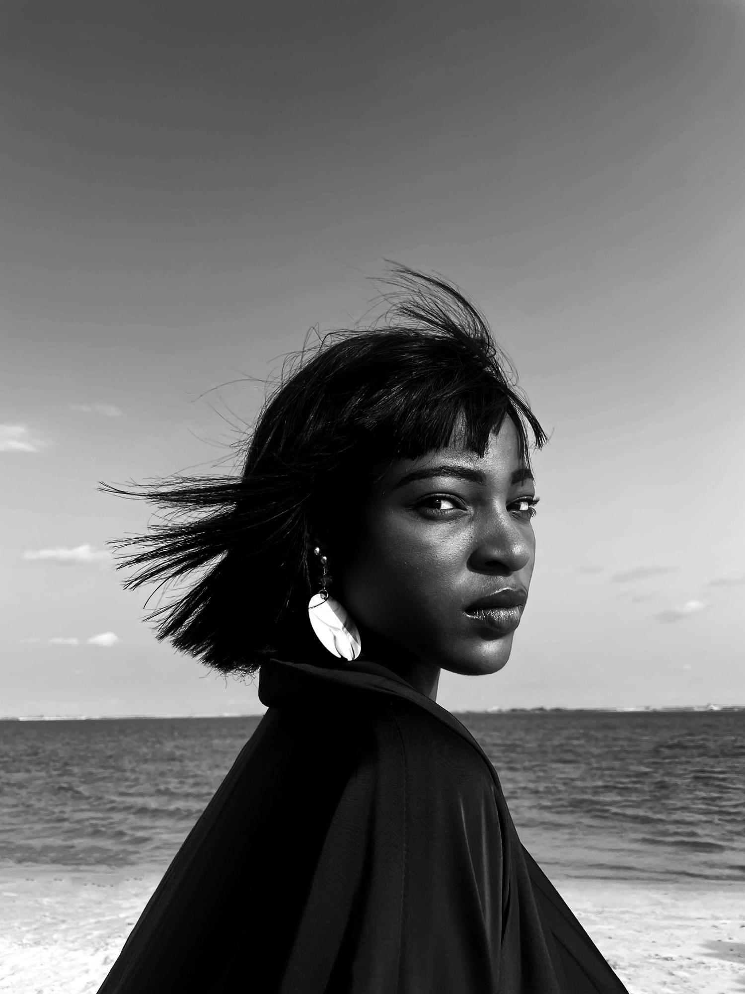 A woman with short hair and a black jacket standing on a beach, looking out at the ocean.