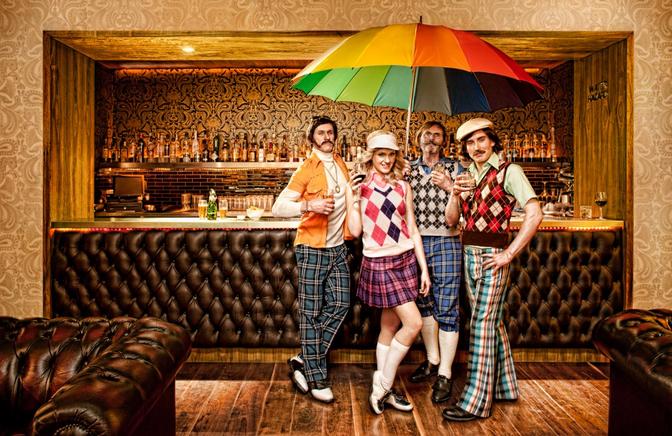 Four people, three men and one woman, dressed in golfing attire, including golfing hats, golfing shirts, golfing pants, and golfing shoes, standing in a bar, holding drinks and smiling.