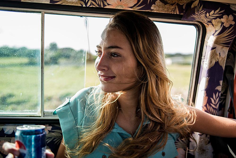 A young woman with long blonde hair and a blue shirt is sitting in a car, looking out the window at a green field.
