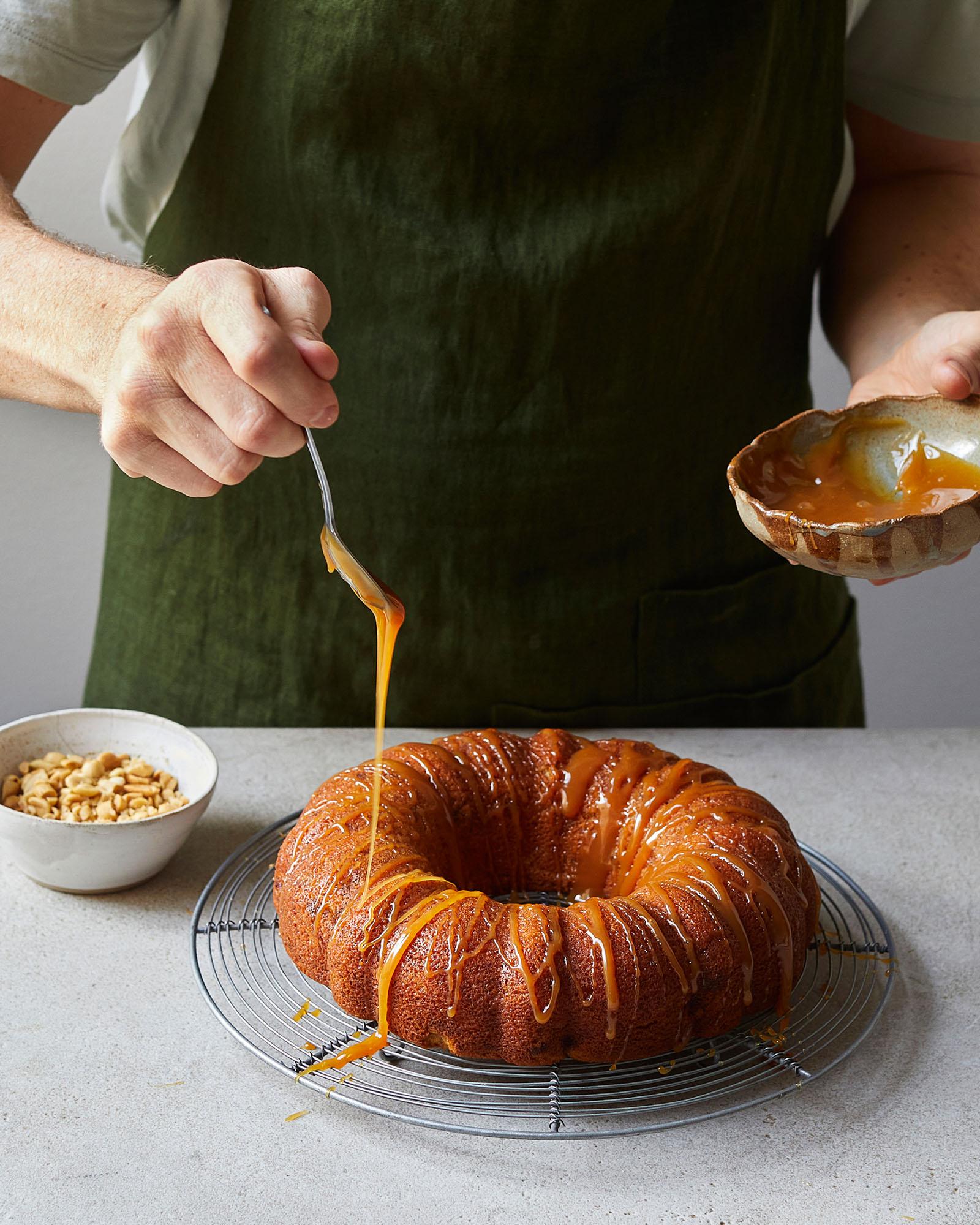 A person in a green apron is drizzling a sweet, golden sauce over a ring-shaped cake.