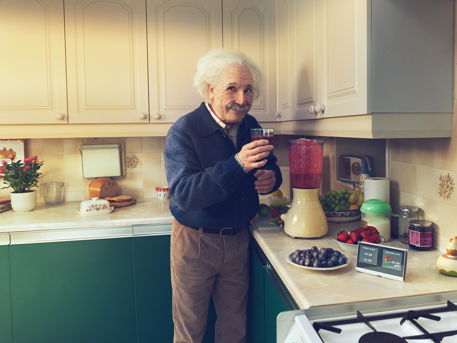 A man with white hair and a mustache, wearing a blue sweater and holding a glass of red liquid, standing in a kitchen with a blender, fruit, and a stove.