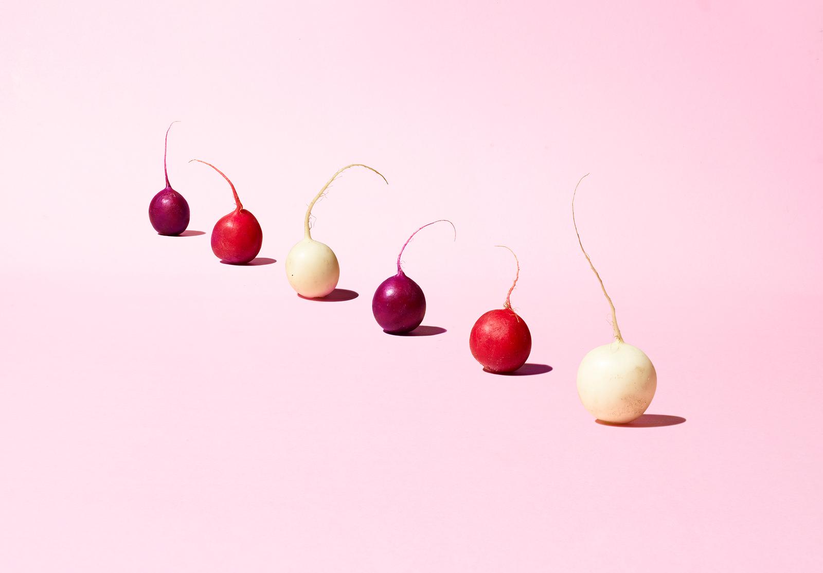 A group of radishes in various colors, including red, white, and purple, arranged in a line on a white background.